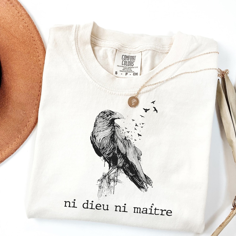 May include: Cream-colored t-shirt featuring a detailed black raven illustration with the phrase "ni dieu ni ma&icirc;tre" printed below. A gold necklace and a brown hat are also visible. The shirt is made of Comfort Colors material.