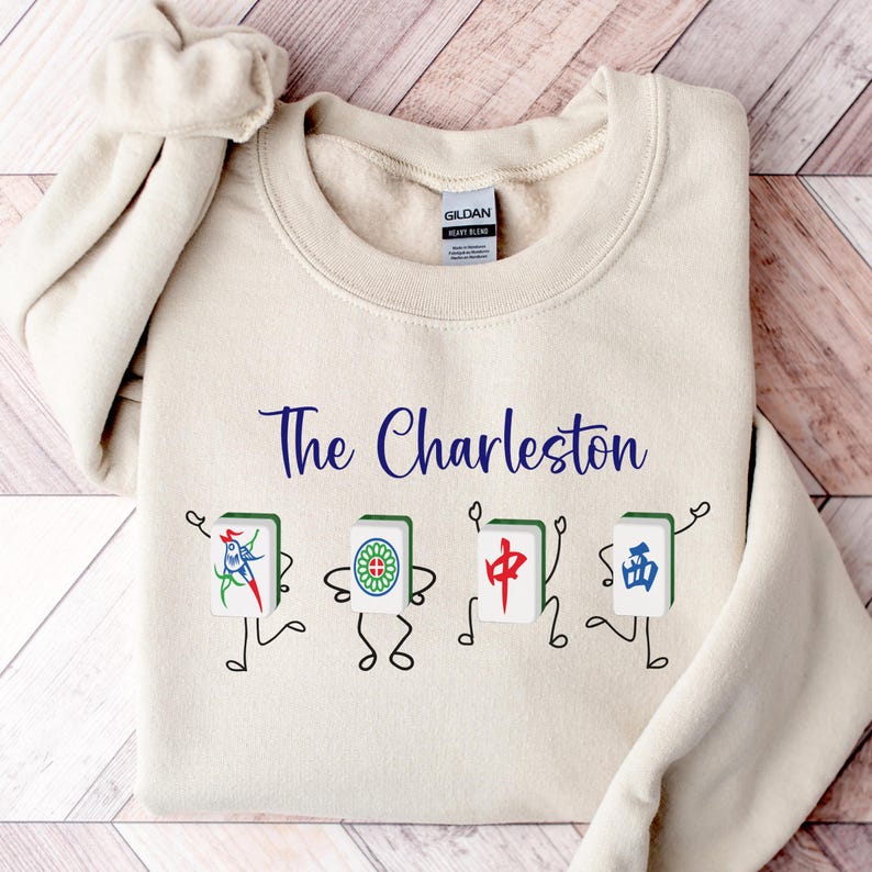 May include: A beige sweatshirt with the text "The Charleston" printed in blue. Below the text are four cartoon characters holding mahjong tiles with Chinese characters.