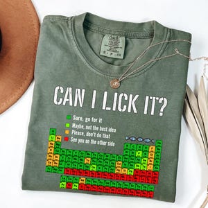 May include: Olive green t-shirt with the text "CAN I LICK IT?" and a periodic table design. The shirt also has a list of options with corresponding colors. A silver necklace with a pendant is draped across the shirt.
