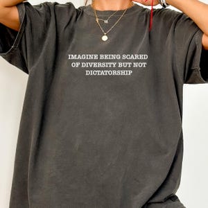 May include: Dark gray t-shirt with the text "IMAGINE BEING SCARED OF DIVERSITY BUT NOT DICTATORSHIP" in white. The shirt has a relaxed fit and short sleeves. The image shows the top of the shirt.