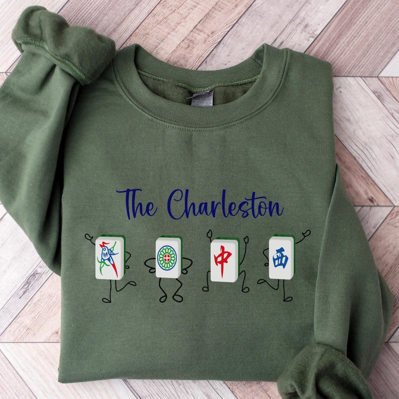 May include: A green sweatshirt with the text "The Charleston" printed on the front. The sweatshirt features a design of four cartoon mahjong tiles with stick figures.
