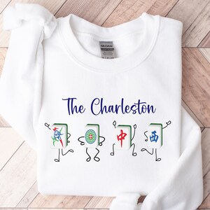 May include: White sweatshirt with blue text that reads "The Charleston" and a graphic of four cartoon mahjong tiles with Chinese characters.