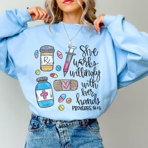 Könnte beinhalten: Hellblauer Pullover mit einem grafischen Design einer Spritze, Pillen und einem Verband. Der Text lautet "She works willingly with her hands Proverbs 31:13".