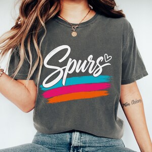 May include: A charcoal grey short-sleeve t-shirt featuring the word "Spurs" in white cursive script with a small heart icon. Below the text are three horizontal brushstroke stripes in bright turquoise, hot pink, and vibrant orange colours.