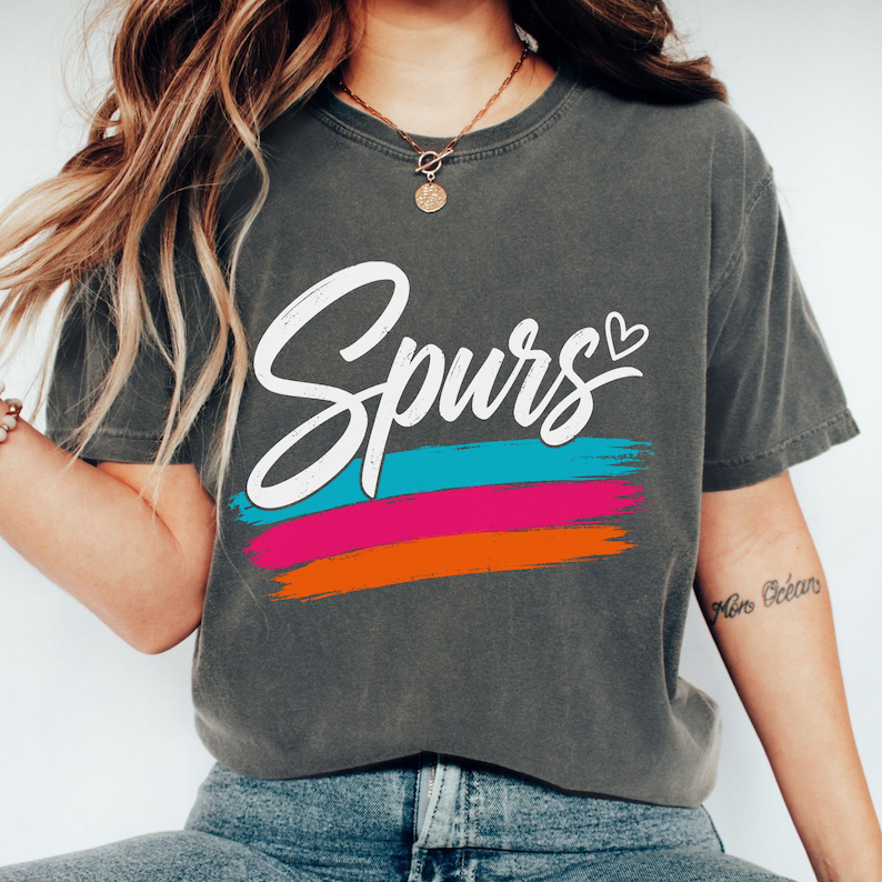 May include: A charcoal grey short-sleeve t-shirt featuring the word "Spurs" in white cursive script with a small heart icon. Below the text are three horizontal brushstroke stripes in bright turquoise, hot pink, and vibrant orange colours.