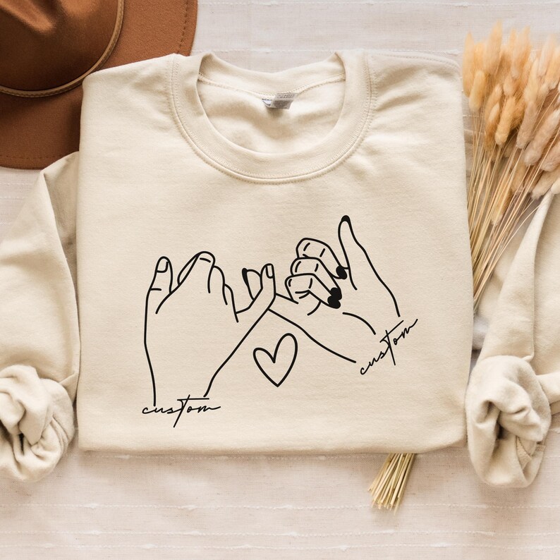 Puede incluir: Una sudadera beige con un contorno negro de dos manos haciendo una promesa me&ntilde;ique. El texto "custom" est&aacute; escrito debajo de cada mano.