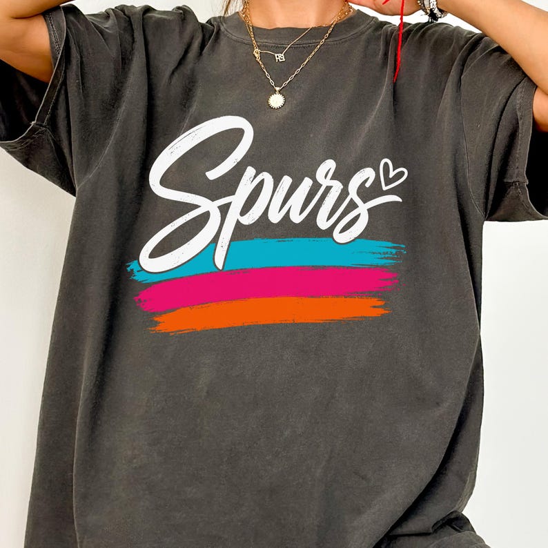 May include: A charcoal grey short-sleeve t-shirt featuring the word Spurs in white cursive script with a small heart icon. Below the text are three horizontal brushstroke stripes in bright turquoise, hot pink, and vibrant orange colours.