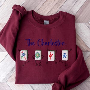 May include: Burgundy sweatshirt with the text "The Charleston" in blue lettering and a graphic of four cartoon figures holding mahjong tiles.