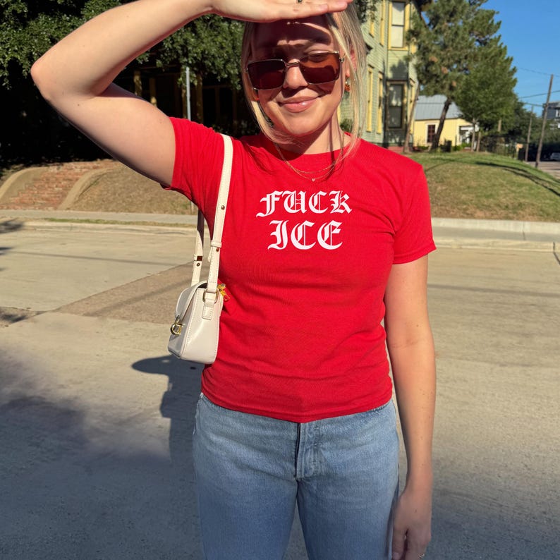Pu&ograve; includere: Una maglietta rossa con la scritta "FUCK ICE" in lettere gotiche bianche. La maglietta &egrave; abbinata a jeans blu chiaro e una piccola borsa bianca sporca. La persona indossa occhiali da sole all'aperto in una giornata di sole.