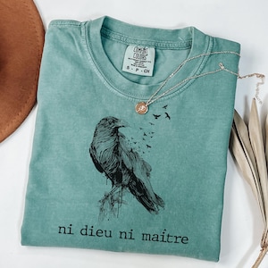 Könnte beinhalten: Ein gefaltetes, meergrünes T-Shirt mit einem schwarzen Raben-Design und dem französischen Text "ni dieu ni maître". Das Shirt hat ein Comfort Colors-Etikett. Eine silberne Halskette mit Anhänger und Vögeln liegt darauf.