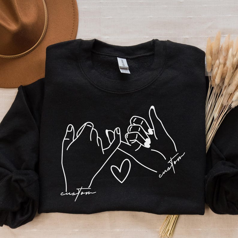 Puede incluir: Sudadera negra con un contorno blanco de dos manos que se agarran los me&ntilde;iques, con un coraz&oacute;n en el medio y la palabra "custom" escrita debajo de cada mano.