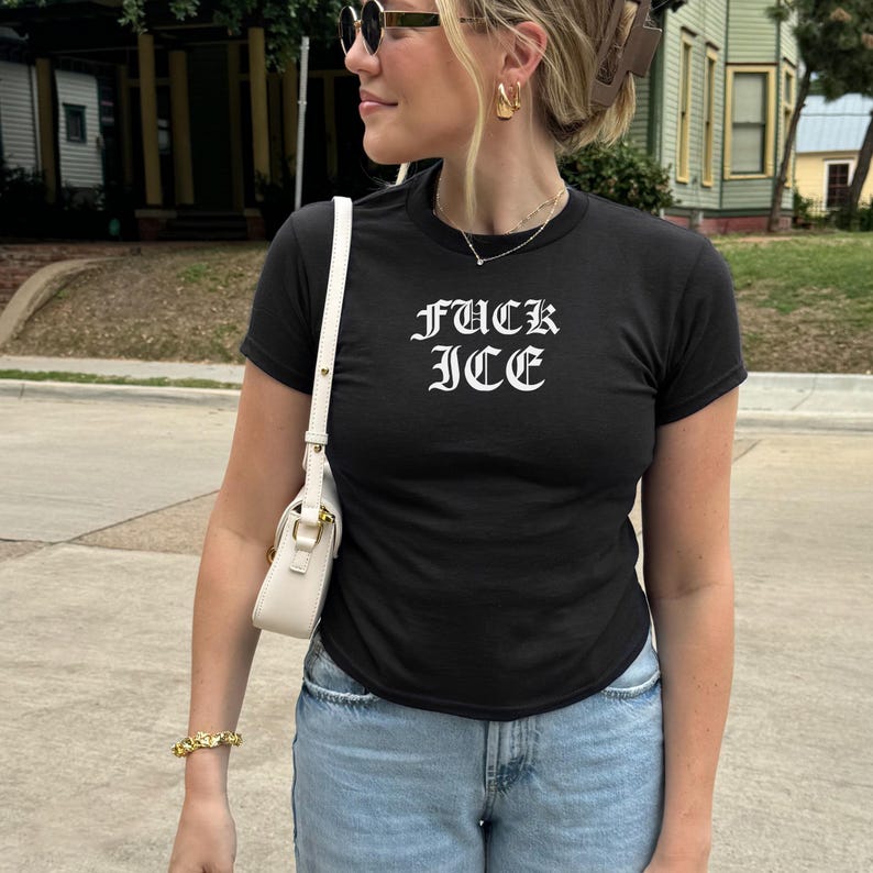 Pu&ograve; includere: T-shirt nero girocollo con la scritta "FUCK ICE" in carattere gotico. La persona indossa jeans blu, occhiali da sole e una borsa bianca. Sono visibili anche gioielli dorati, tra cui un braccialetto e orecchini.