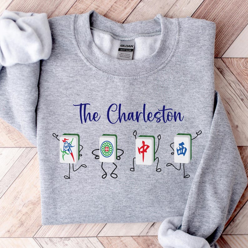 May include: Gray sweatshirt with the text "The Charleston" printed in blue cursive font. Four cartoon mahjong tiles are arranged below the text, each with a different symbol and a pair of legs.