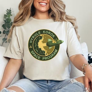 Peut inclure: Un t-shirt beige avec un graphique circulaire vert et jaune représentant une fusée décollant. Le graphique indique "Hyrule Korok Space Program".
