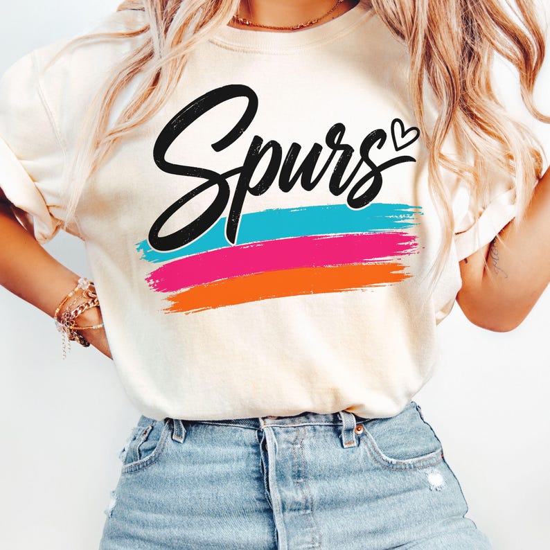 May include: A cream-coloured short-sleeve t-shirt featuring the word Spurs in black cursive script with a small heart above the letter s. Below the text are three horizontal brushstroke stripes in bright turquoise, hot pink, and orange colours.