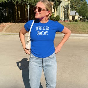 Pu&ograve; includere: Una maglietta blu reale con la scritta "FUCK ICE" in lettere gotiche bianche. La maglietta &egrave; abbinata a jeans di denim chiaro. Una borsa bianca &egrave; indossata a tracolla.