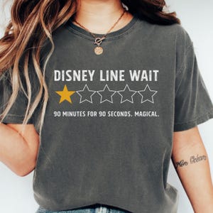 May include: Dark gray t-shirt with the text "DISNEY LINE WAIT" and a rating of one gold star and four outlined stars. The text "90 MINUTES FOR 90 SECONDS. MAGICAL." is below the stars.