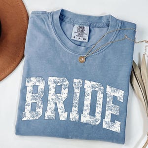 May include: A blue Comfort Colors t-shirt with the word "BRIDE" in large, white, floral-patterned letters. A gold necklace with a pendant rests on the shirt. The shirt is folded and displayed with a brown hat and dried leaves.