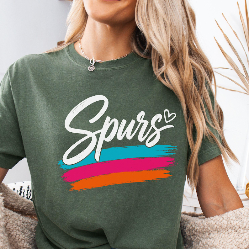 May include: An olive green crew neck t-shirt featuring the word "Spurs" in white cursive script with a small heart icon. Below the text are three horizontal brushstroke stripes in bright turquoise, hot pink, and vibrant orange colours.