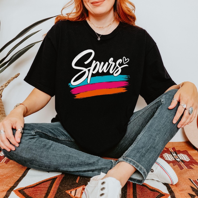 May include: A black short-sleeved t-shirt featuring the word "Spurs" in white cursive script with a small heart above the letter s. Below the text are three horizontal brushstroke lines in teal, magenta, and orange colours on the chest area.