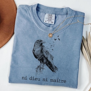 May include: A blue Comfort Colors t-shirt featuring a black raven illustration with the text "ni dieu ni ma&icirc;tre". A gold necklace with a pendant is draped across the shirt. The shirt is folded and placed on a white surface.