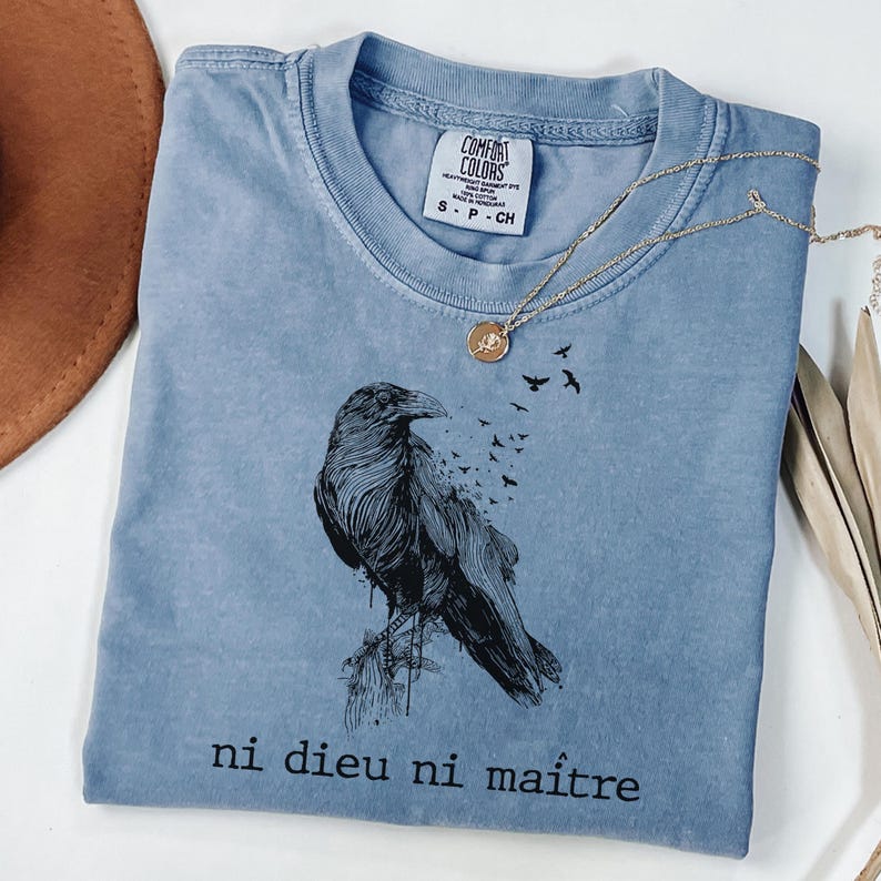May include: A blue Comfort Colors t-shirt featuring a black raven illustration with the text "ni dieu ni ma&icirc;tre". A gold necklace with a pendant is draped across the shirt. The shirt is folded and placed on a white surface.