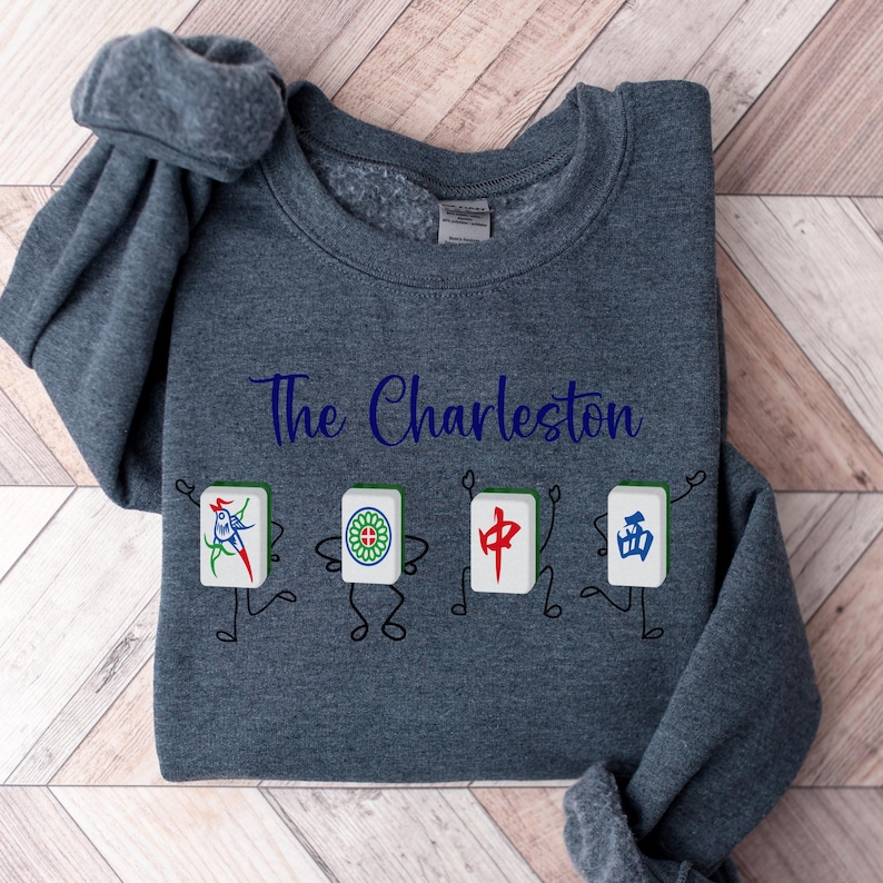 May include: A dark grey sweatshirt with the text "The Charleston" printed in blue. Four cartoon figures are holding mahjong tiles with Chinese characters.