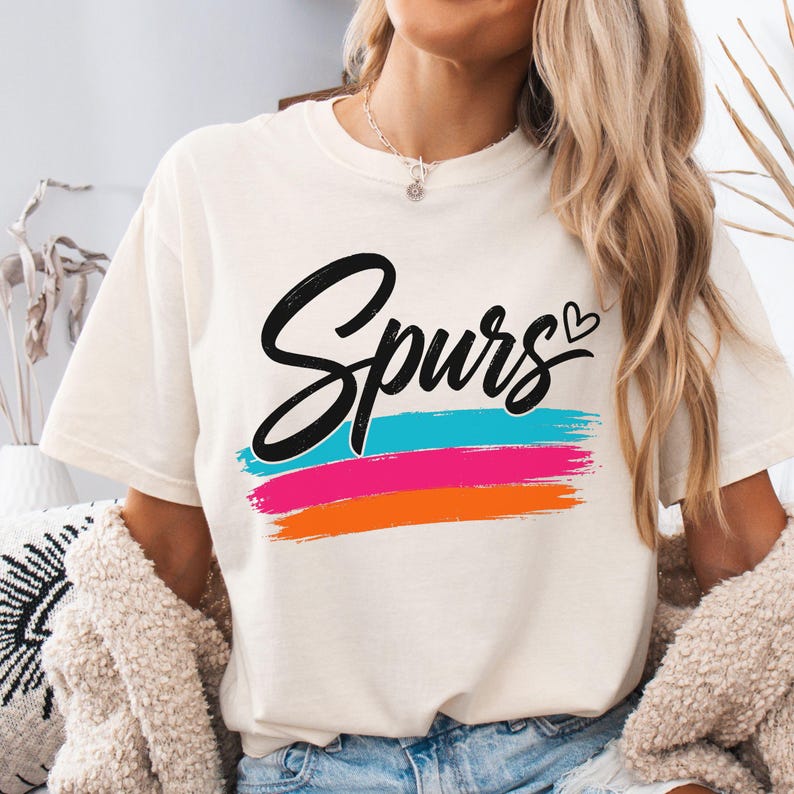 May include: A cream-coloured short-sleeve t-shirt featuring the word Spurs in black cursive script with a small heart icon. Below the text are three horizontal brushstroke stripes in bright turquoise, hot pink, and vibrant orange colours.