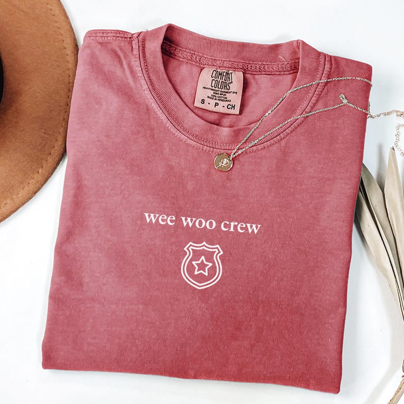 Puede incluir: Una camiseta Comfort Colors rosa descolorido con las palabras "wee woo crew" y un gr&aacute;fico de insignia de estrella. Un collar plateado con un colgante redondo descansa sobre la camisa. Un sombrero marr&oacute;n es visible.