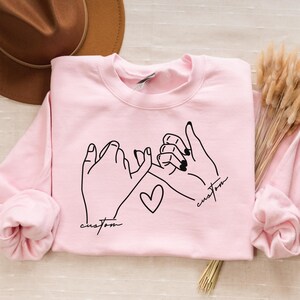 Puede incluir: Sudadera rosa con un contorno negro de dos manos haciendo una promesa me&ntilde;ique con un coraz&oacute;n en el medio. El texto "custom" est&aacute; escrito a cada lado del coraz&oacute;n.