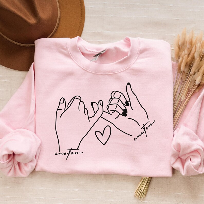 Puede incluir: Sudadera rosa con un contorno negro de dos manos haciendo una promesa me&ntilde;ique con un coraz&oacute;n en el medio. El texto "custom" est&aacute; escrito a cada lado del coraz&oacute;n.