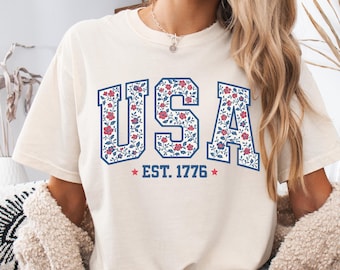 May include: Cream-colored t-shirt with the word "USA" in a blue floral design. The text "EST. 1776" is below the word "USA" with a red star on each side. The shirt has a relaxed fit.