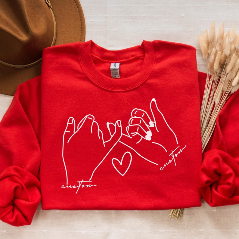 Puede incluir: Sudadera roja con un contorno blanco de dos manos haciendo una promesa me&ntilde;ique con un coraz&oacute;n en el medio. El texto "custom" est&aacute; escrito debajo de cada mano.