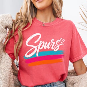 May include: A coral-coloured short-sleeve t-shirt featuring the word Spurs in white cursive script with a small heart icon. Below the text are three horizontal brushstroke stripes in turquoise, hot pink, and orange colours on the front chest.