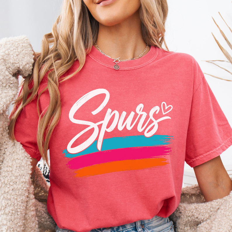 May include: A coral-coloured short-sleeve t-shirt featuring the word Spurs in white cursive script with a small heart icon. Below the text are three horizontal brushstroke stripes in turquoise, hot pink, and orange colours on the front chest.