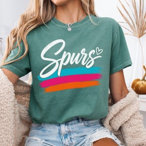 May include: A green short-sleeved t-shirt featuring the word "Spurs" in white cursive script with a small heart icon. Below the text are three horizontal brushstroke stripes in teal, magenta, and orange. The shirt has a crew neckline.