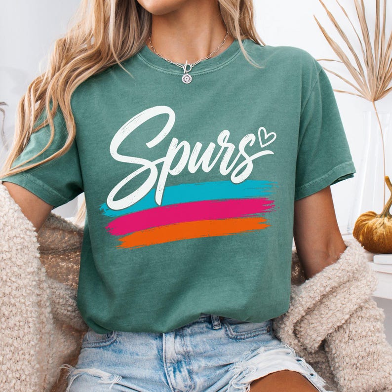 May include: A green short-sleeved t-shirt featuring the word "Spurs" in white cursive script with a small heart icon. Below the text are three horizontal brushstroke stripes in teal, magenta, and orange. The shirt has a crew neckline.