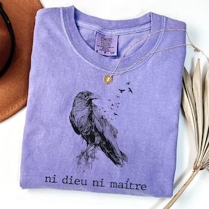 May include: A lavender Comfort Colors t-shirt featuring a detailed black raven illustration with the phrase "ni dieu ni ma&icirc;tre" printed below. A gold necklace with a pendant rests on the shirt.