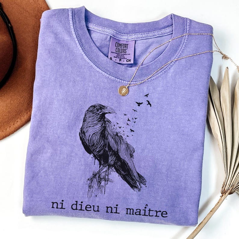May include: A lavender Comfort Colors t-shirt featuring a detailed black raven illustration with the phrase "ni dieu ni ma&icirc;tre" printed below. A gold necklace with a pendant rests on the shirt.
