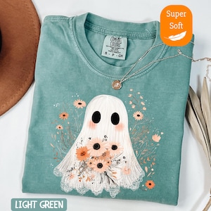 May include: Light green t-shirt featuring a ghost illustration holding a bouquet of flowers. The ghost has large black eyes and blush-colored cheeks. The shirt has a crew neck and short sleeves. The text "LIGHT GREEN" is at the bottom of the shirt.