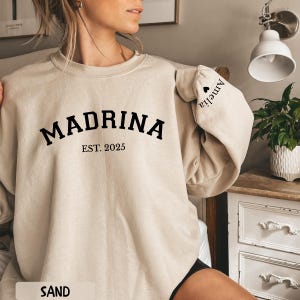 May include: Sand-colored sweatshirt with the word "MADRINA" in bold black letters, with "EST. 2025" underneath. The sleeve has the name "Amelia" printed on it. The word "SAND" is at the bottom of the image.