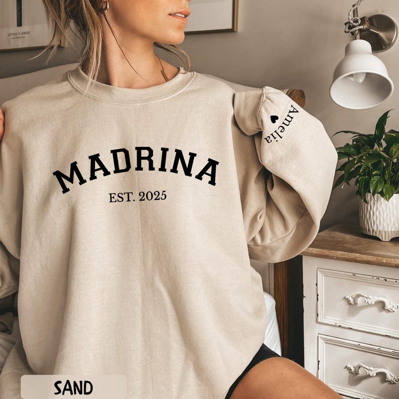 May include: Sand-colored sweatshirt with the word "MADRINA" in bold black letters, with "EST. 2025" underneath. The sleeve has the name "Amelia" printed on it. The word "SAND" is at the bottom of the image.