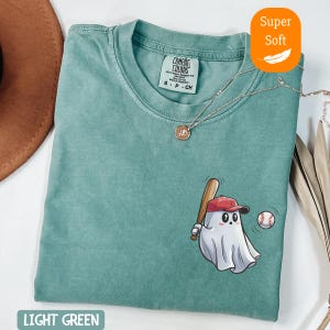 May include: Light green t-shirt with a baseball-themed ghost graphic. The ghost wears a red baseball cap and holds a bat, with a baseball nearby. The shirt has a crew neck and a small tag.