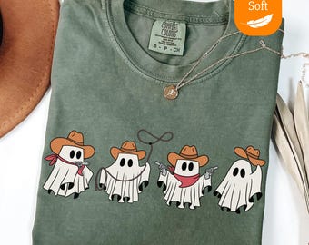 May include: A moss green t-shirt featuring a graphic of four cartoon ghosts dressed as cowboys, complete with hats, bandanas, and toy guns. The shirt has a crew neck and short sleeves. A gold necklace is draped across the shirt.