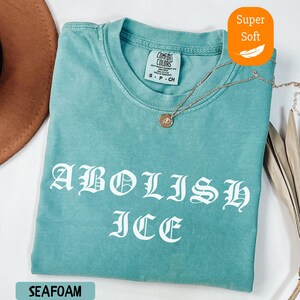 May include: Seafoam green t-shirt with the words "ABOLISH ICE" in white gothic lettering. The shirt has a crew neck and short sleeves. A gold necklace with a pendant rests on the shirt. The tag reads "COMFORT COLORS".