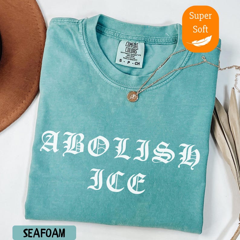 May include: Seafoam green t-shirt with the words "ABOLISH ICE" in white gothic lettering. The shirt has a crew neck and short sleeves. A gold necklace with a pendant rests on the shirt. The tag reads "COMFORT COLORS".