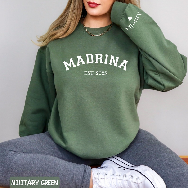 May include: Military green sweatshirt with the word "MADRINA" in white block letters, with "EST. 2025" below. The sleeve has the name "Amelia" printed in white. The sweatshirt is a crew neck style.