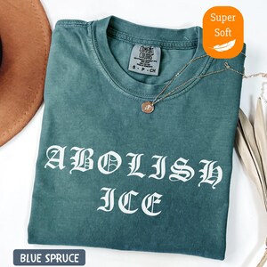 May include: Blue spruce-colored t-shirt with the words "ABOLISH ICE" in white gothic lettering. The shirt has a crew neck and short sleeves. A gold necklace with a pendant is draped on the shirt.