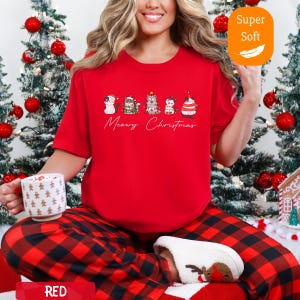 May include: A red t-shirt with the text "Meowy Christmas" and a row of cartoon cats in festive outfits. The shirt is paired with red and black plaid pajama pants. A white mug with a star pattern is held in the hand.