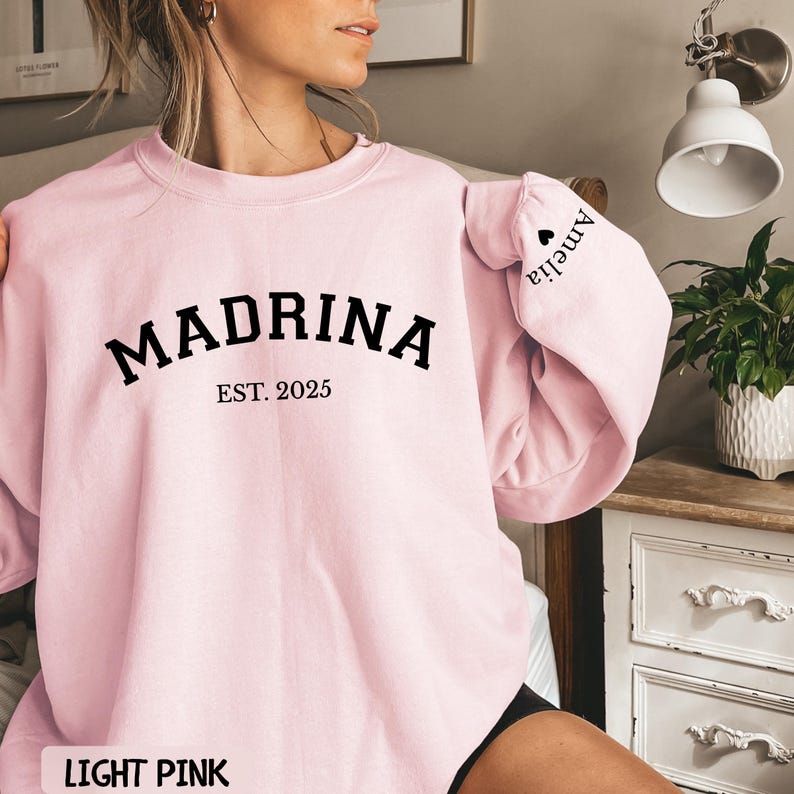 May include: Light pink sweatshirt with the word "MADRINA" in bold black letters, with "EST. 2025" underneath. The sleeve has the name "Amelia" printed on it. The bottom of the sweatshirt says "LIGHT PINK".